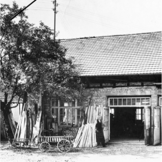 Historische Werkstatt mit offenen Türen, Holzlatten an der Wand und einem Wagen davor. Eine Person steht in der Tür.