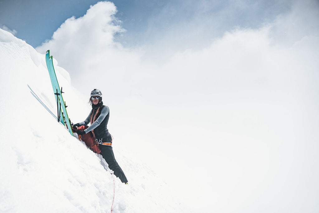 Eine Frau mit Kletterausrüstung steht an einem steilen, verschneiten Hang, während ihre Skier senkrecht neben ihr in den Schnee gesteckt sind