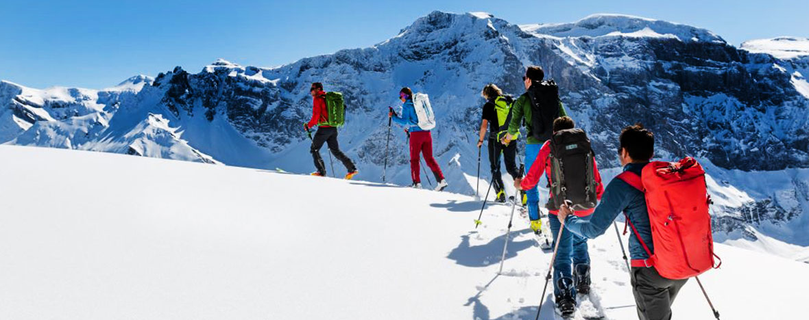 Eine Gruppe von Skitourengehern geht in einer Linie über einen unberührten Schneehang, mit dramatischen Berglandschaften im Hintergrund.