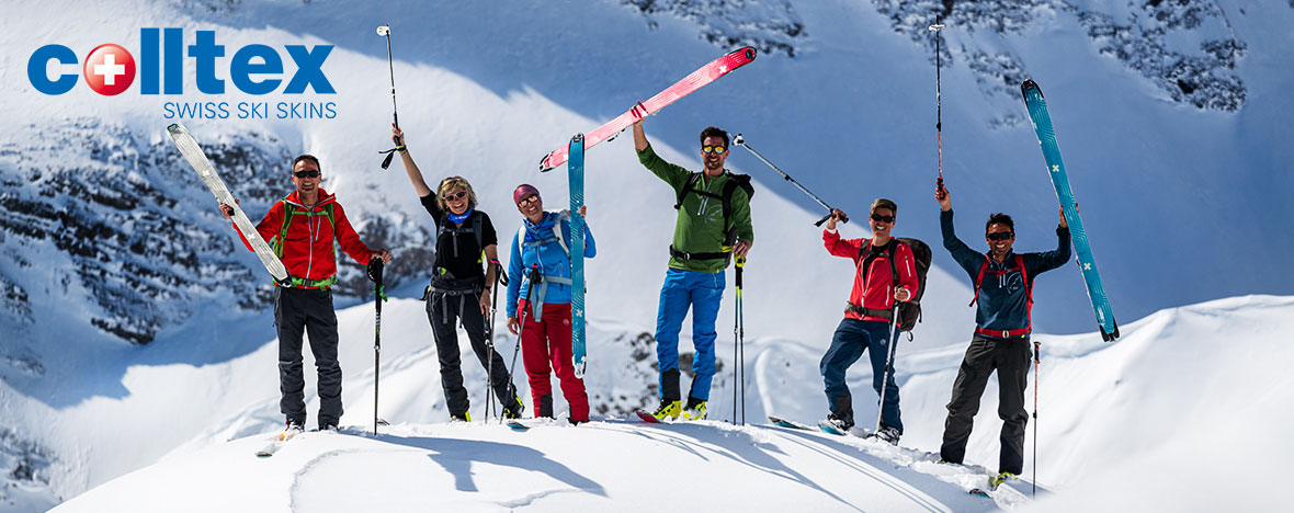 Eine Gruppe von Skifahrern posiert auf einem schneebedeckten Gipfel, umgeben von schroffen Berggipfeln, mit Skiern und Skistöcken in der Hand.