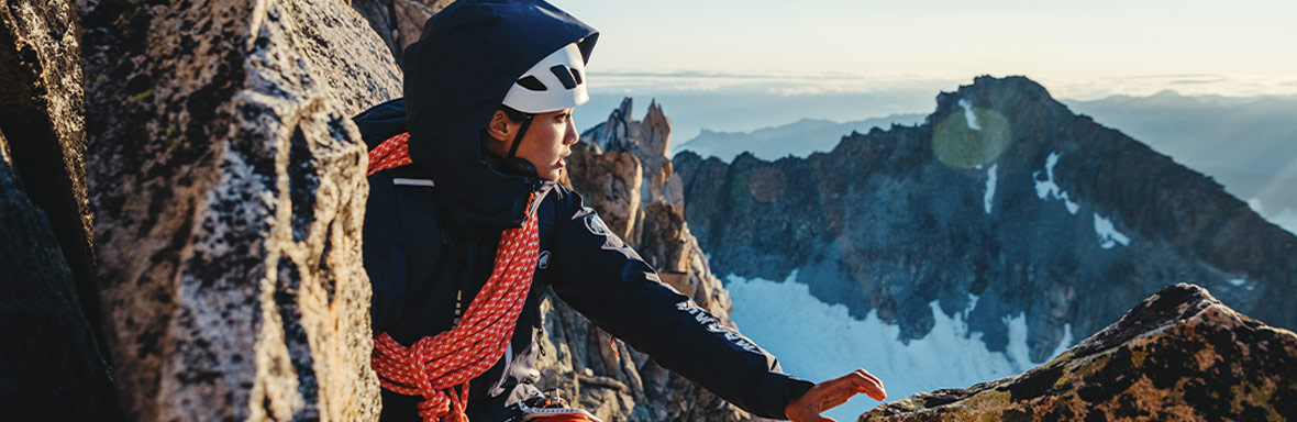 Ein Bergsteiger mit dunkler Ausrüstung und weißem Helm, der eine orangefarbene Kletterseilschlinge trägt und auf einer felsigen Anhöhe sitzt. Der Hintergrund zeigt eine beeindruckende Berglandschaft mit schneebedeckten Gipfeln bei Sonnenaufgang oder Sonnenuntergang.