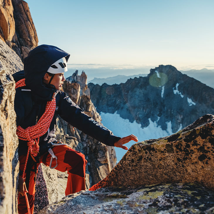 Ein Bergsteiger mit dunkler Ausrüstung und weißem Helm, der eine orangefarbene Kletterseilschlinge trägt und auf einer felsigen Anhöhe sitzt. Der Hintergrund zeigt eine beeindruckende Berglandschaft mit schneebedeckten Gipfeln bei Sonnenaufgang oder Sonnenuntergang.