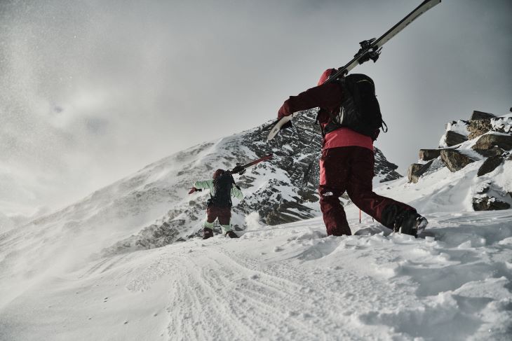 Zwei Skifahrer in roter Ausrüstung tragen ihre Ski einen verschneiten Berghang hinauf