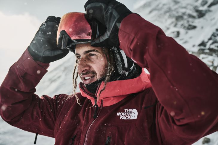 Nahaufnahme eines Skifahrers in roter Jacke, der seine Skibrille aufsetzt, im Hintergrund verschneite Berge