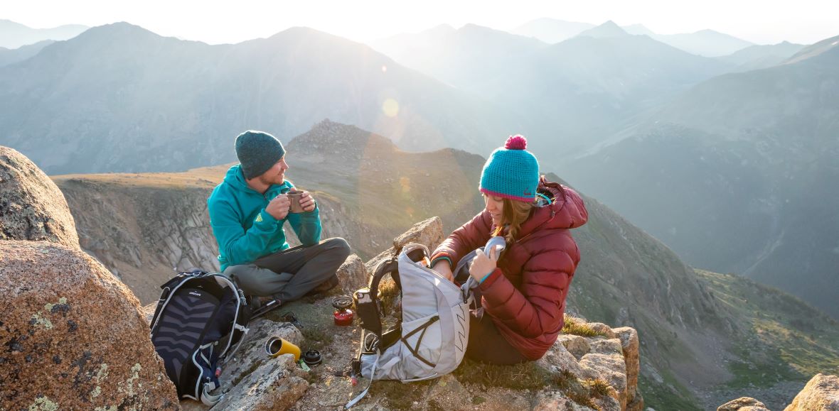 Zwei Wanderer sitzen auf einem felsigen Gipfel, genießen eine Pause und trinken heiße Getränke, während die Morgensonne über die Berglandschaft scheint.