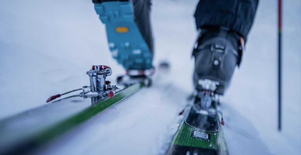 Nahaufnahme einer Skibindung und eines Skistiefels auf einer grünen Skikante, die Szene zeigt einen Skifahrer in Bewegung auf frischem Schnee.