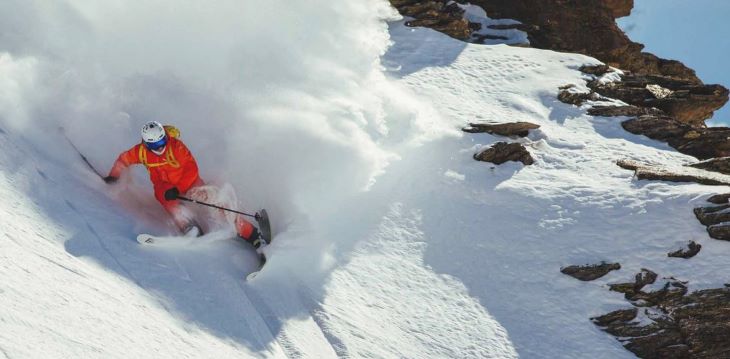 Ein Skifahrer in einer roten Skijacke fährt einen steilen Hang hinunter, umgeben von aufwirbelndem Schnee, der ihn in einer dynamischen Bewegung zeigt.