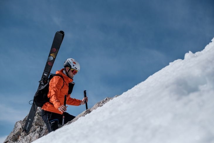 Ein Bergsteiger in einer orangefarbenen Jacke und Helm trägt seine Skier auf dem Rücken und klettert einen schneebedeckten Hang hinauf. Der Himmel ist strahlend blau und verleiht der Szene eine dramatische Note.