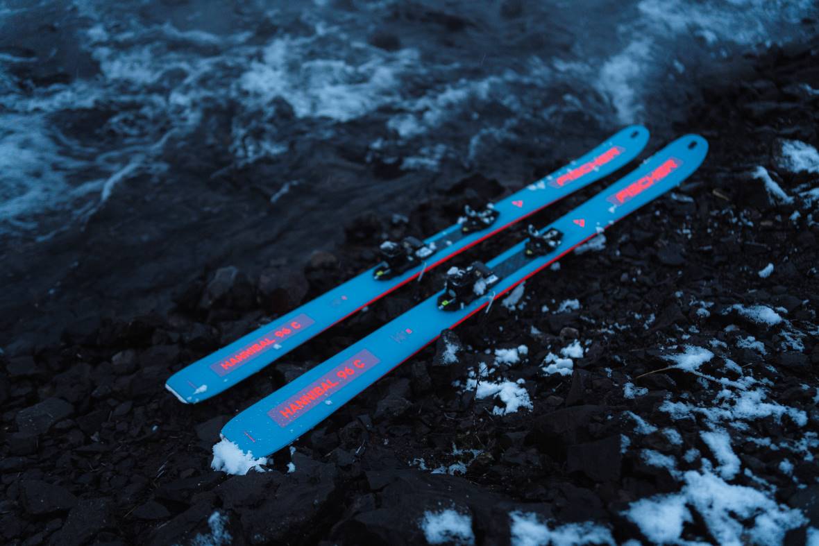 Ein Paar Fischer-Ski mit der Aufschrift 'Hannibal 96 C' liegt auf einer mit etwas Schnee bedeckten steinigen Oberfläche neben einem Fluss oder See.