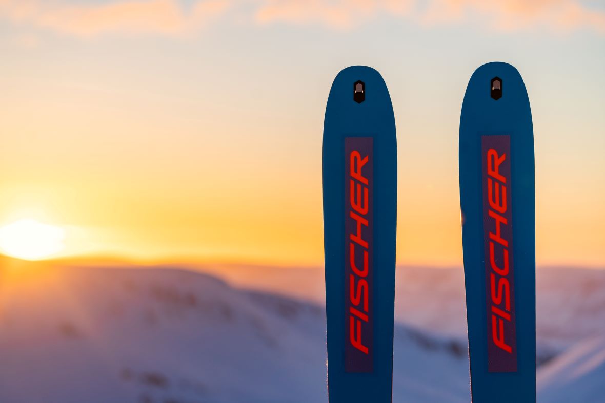 Zwei blaue Skispitzen mit dem roten Aufdruck 'Fischer', die bei Sonnenuntergang in den Himmel ragen. Im Hintergrund sind verschwommene Berge zu sehen.