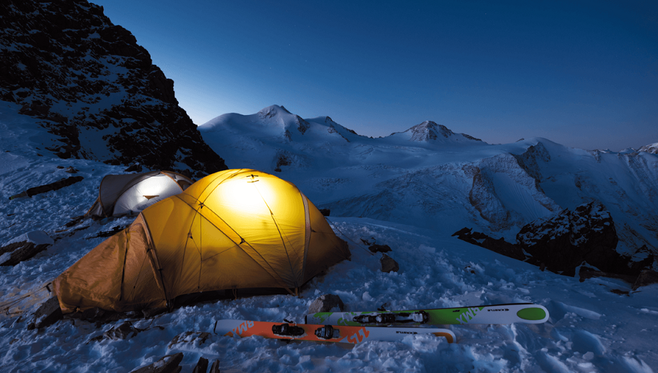 Ein beleuchtetes Zelt steht in der Dämmerung auf einem schneebedeckten Bergplateau, umgeben von weiteren Zelten und Skiausrüstung. Im Hintergrund sind majestätische, verschneite Berggipfel unter dem sternenbedeckten Nachthimmel zu sehen.