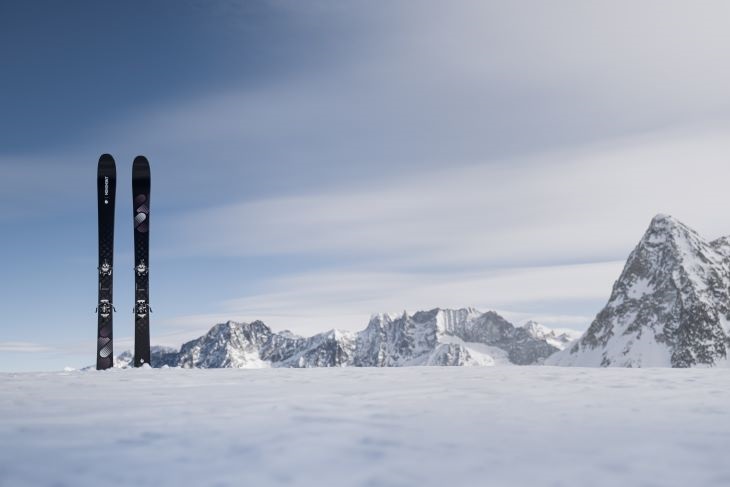 Zwei schwarze Skier stehen im tiefen Schnee vor einer malerischen Berglandschaft. Der Hintergrund zeigt eine beeindruckende Bergkette unter einem klaren Himmel, was die Weite und Schönheit der alpinen Umgebung betont.