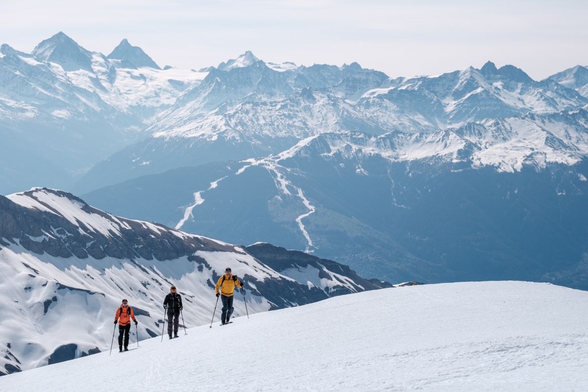Drei Personen wandern im Aufstieg auf einer schneebedeckten Berghangfläche, mit einer atemberaubenden Berglandschaft im Hintergrund, die von Schnee und markanten Gipfeln geprägt ist.