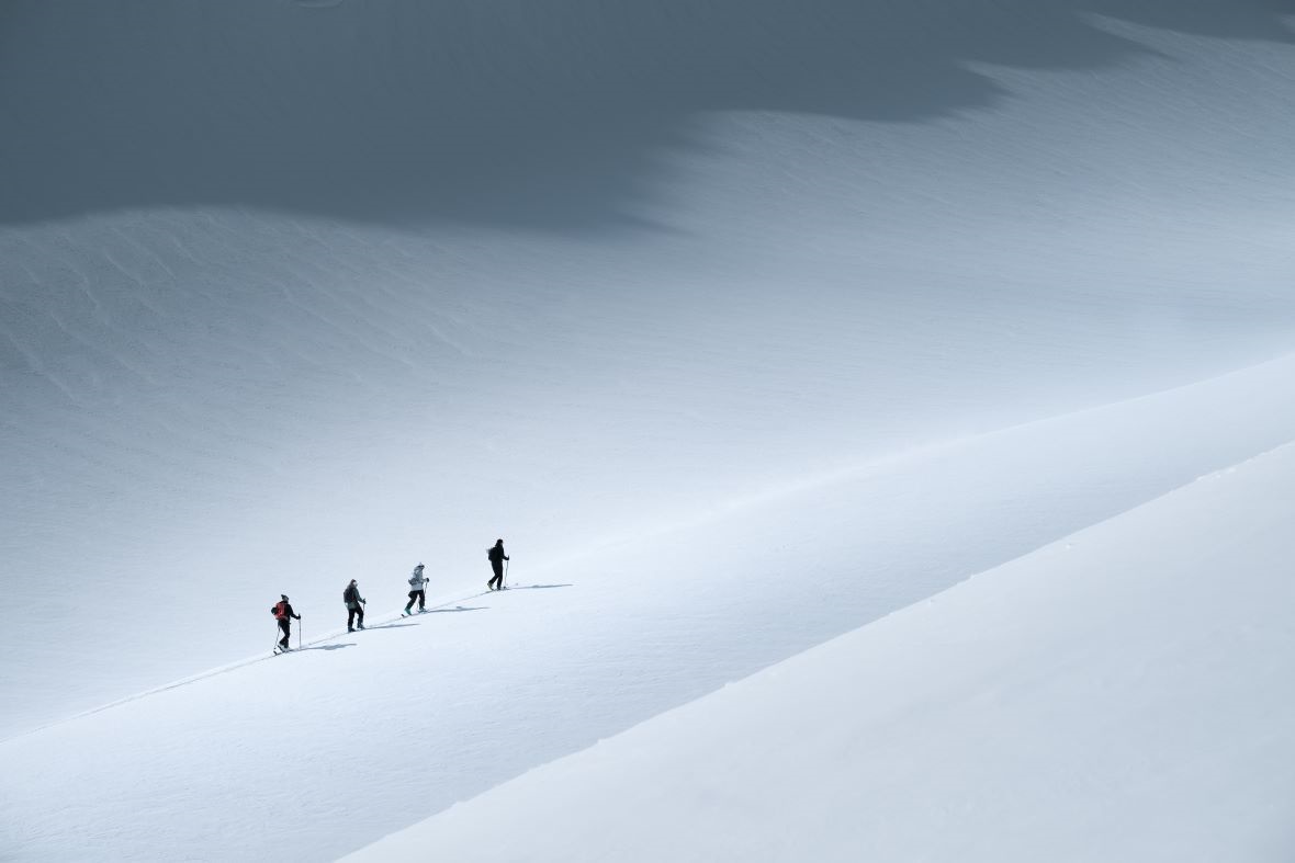 Vier Personen in einer geordneten Reihe beim Skitourenaufstieg auf einem schneeweißen, weiten Hang, unter dramatischen Licht- und Schattenspielen auf der Schneedecke.